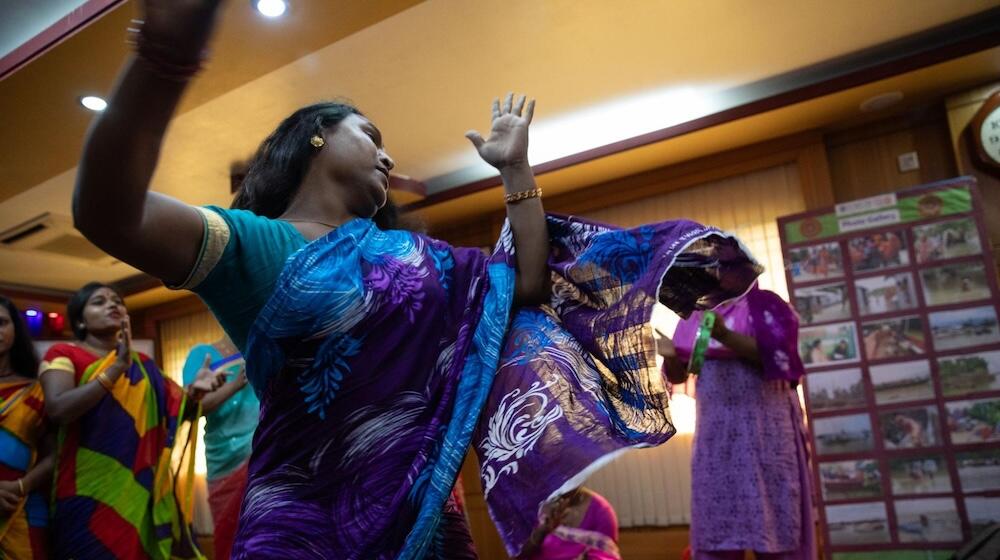A group of transgender and gender-diverse people wearing colourful clothing dance and clap.