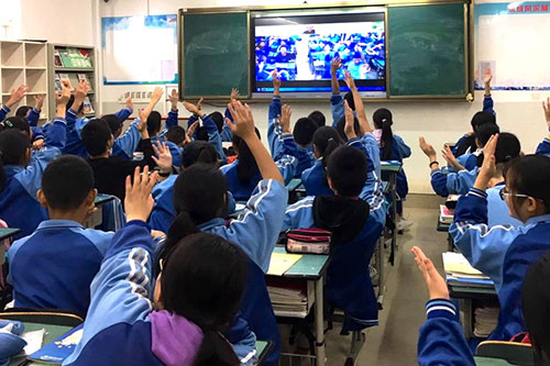 Students raise their hands, interacting with a course on the large screen in their classroom.