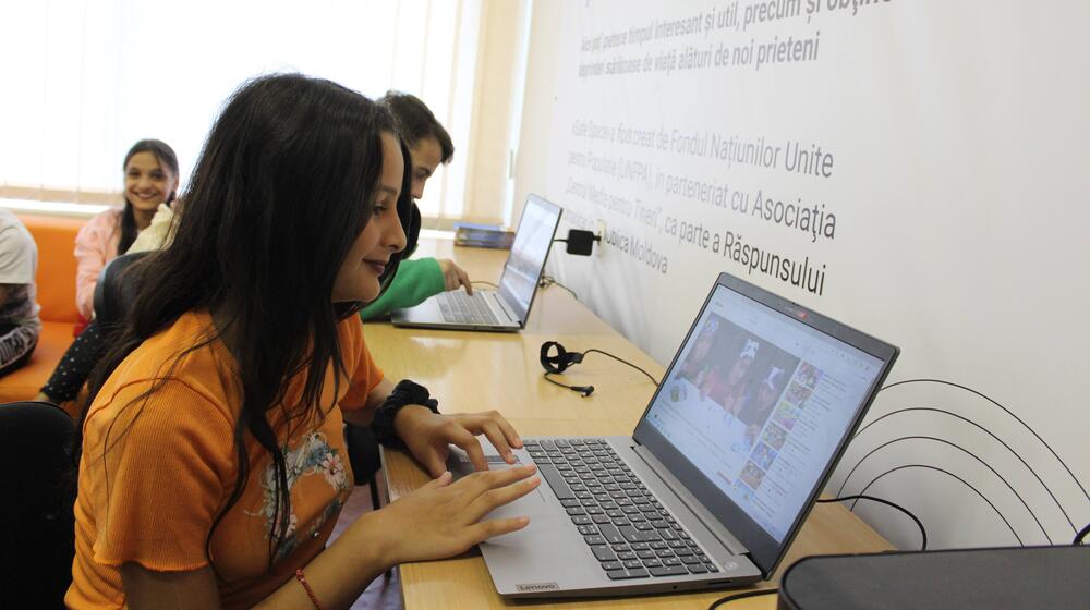 A young woman in an orange t-shirt watches a video on a laptop.