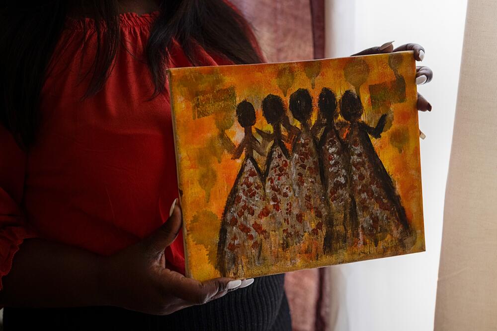 A woman holds up an abstract painting of several figures against an orange background.