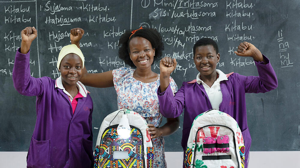 Three young girls cheer with their smart bags, which include a solar panel used to charge a torch to help with their studies when and where there is no electricity.