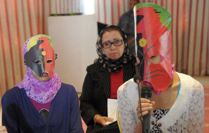 Two adolescent girls are wearing masks and one speaks into a microphone. Behind them stands a moderator.
