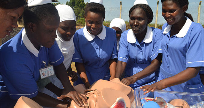 A group of midwives dressed in blue gather and observe from a mannequin