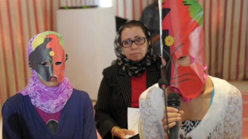 Two adolescent girls are wearing masks and one speaks into a microphone. Behind them stands a moderator.
