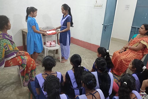 Two girls stand before a crowd of seated adolescent girls. They are holding a clean menstrual pad. 