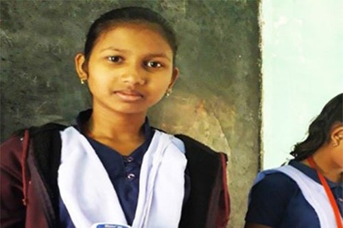 A girl smiles into the camera. She is wearing a purple jacket over her school uniform.