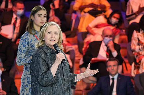 Hillary Clinton holds a microphone in her hand as she delivers remarks. A young activist stands behind her.