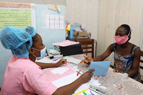 A woman in a pink face mask speaks to a nurse wearing a mask and hair covering.