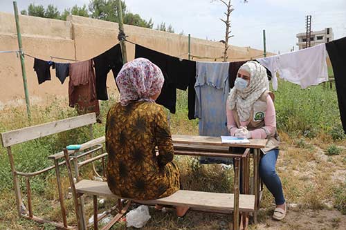 A woman in a face mask speaks with another woman at an outdoor bench.