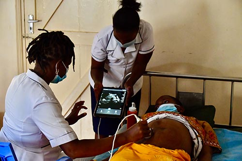 Two midwives in face masks and white uniforms perform an ultrasound on a pregnant woman, who is also wearing a face mask.