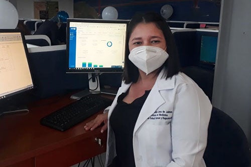 A doctor wearing a face mask sits at a computer with two monitors.