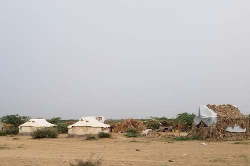 Three tents stand on a sandy ground. One on the left is covered in thatch.