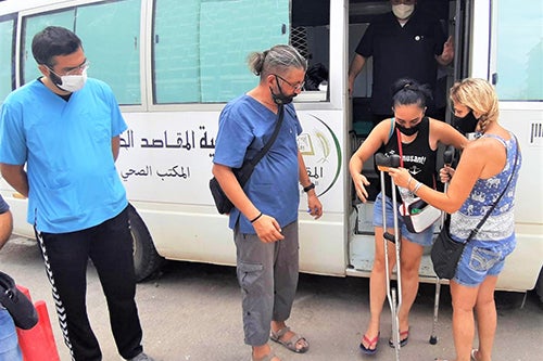 A with a mask hands a crutch to another woman who is limping from the stairs of a mobile clinic.