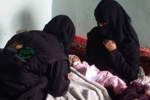 Midwife Lena Al-Shurmani looks at a newborn baby swaddled in a pink blanket. The baby is in her mother's arms. Both the midwife and mother are wearing niqab.