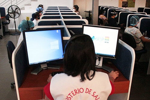 A room with desks and desk dividers is occupied by health workers wearing face masks and hair coverings.