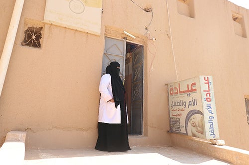 A health worker in a black veil and white lab coat stands outside a clinic.