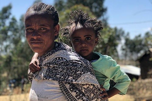 A woman gives a determined look straight into the camera. She is holding a small child on her back.