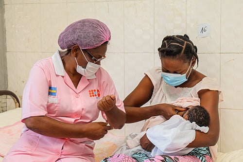A midwife in a face mask and hair cover helps a new mother learn to breastfeed. The new mother is also wearing a facemask.