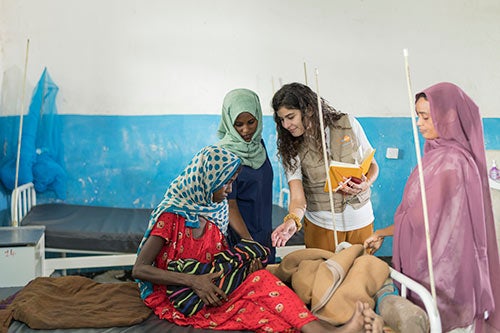 Three women gather around a maternity ward bed, looking at a new baby, held by its mother. 