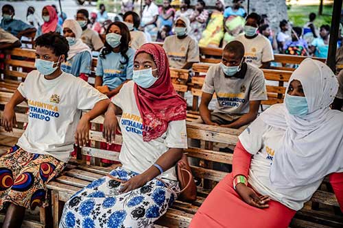 Women sit in an outdoor information session about family planning. They wear colourful skirts, headscarfs and facemasks.