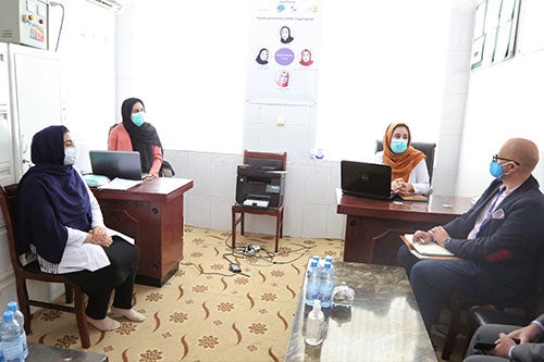 Three women in head scarves and face masks meet with a man wearing a face mask and glasses. 