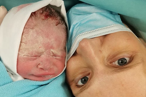 A newborn baby is placed alongside its mother, who is wearing a facemask.