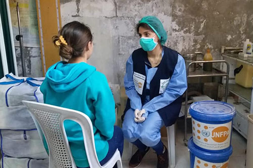 An adolescent girl wearing a blue sweater speaks to a health worker wearing scrubs, a face mask and a hair covering.
