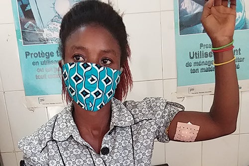 A young woman in a blue face mask raises her arm to show a bandage covering the back of her upper arm.