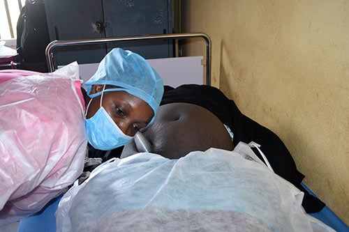 A midwife wearing a mask and hair covering listens to a pregnant woman's abdomen with a pinard stethoscope.