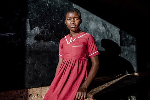 A young girl in a red school dress looks seriously at the camera. Behind her is a chalkboard.