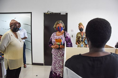 Women and men in face masks stand and listen to a counsellor speak about her experiences. The counsellor's back is to the camera.