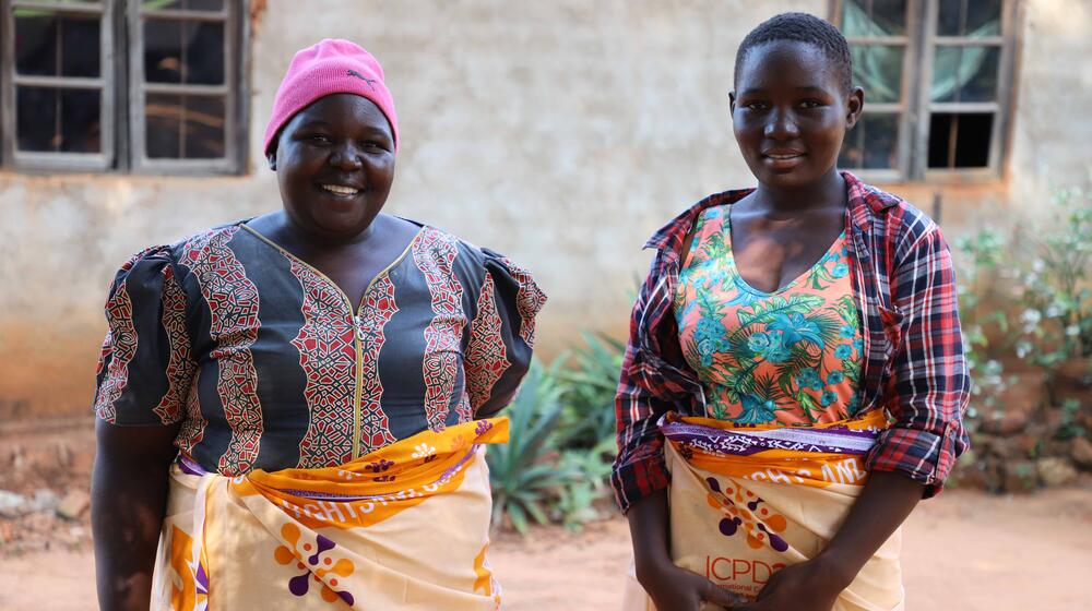 Two women - a mother and daughter - stand together. They both wear bright UNFPA clothing.
