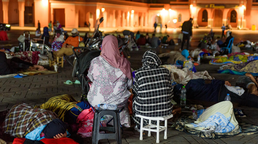 Two people sit on small chairs. They are outside with many other individuals who are sleeping on the ground.