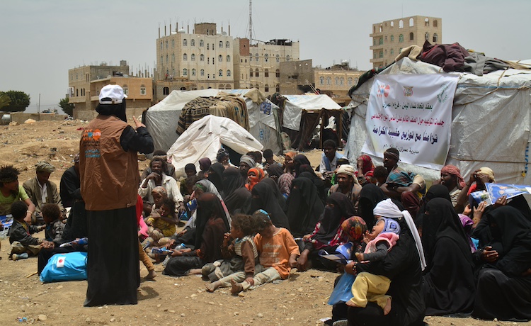  امرأة تقف أمام مجموعة كبيرة من الأشخاص الجالسين على الأرض في مخيم للنازحين وتقدم جلسة حول التوعية بالعنف القائم على النوع الاجتماعي.
