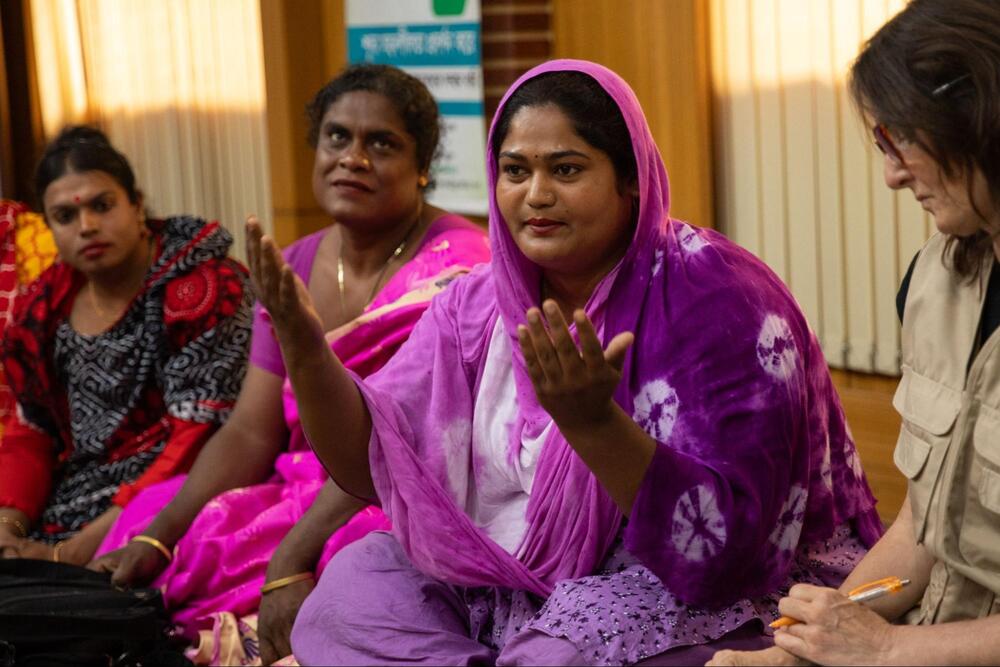 Maimouni, a member of Bangladesh’s hijra group, is dressed in pink and purple clothing and sits and speaks among a group.