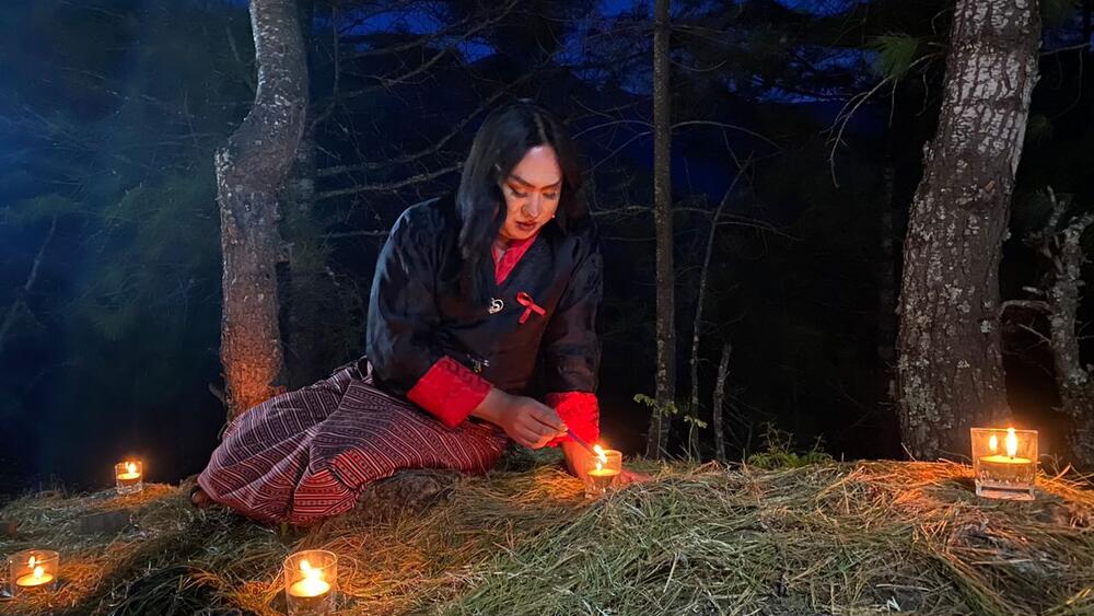 A transgender woman lights a candle against a backdrop of trees at night.