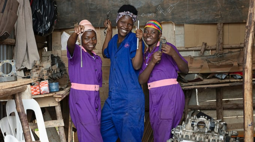Three women wearing protective clothing pose with tools in a mechanical workshop.