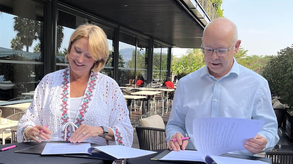 Two individuals - a man and a woman - sit at a table outdoors and sign paperwork.
