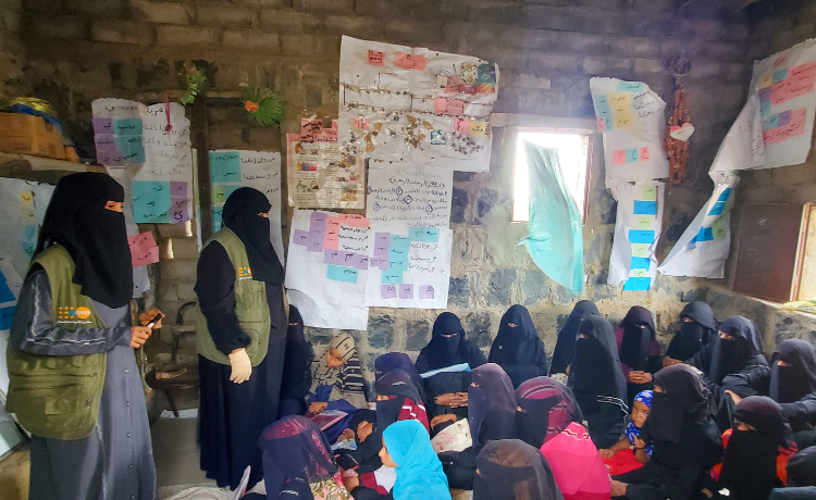 Two women in UNFPA vests and black head covering speak to a group of girls in black head coverings seated on the floor of a small room