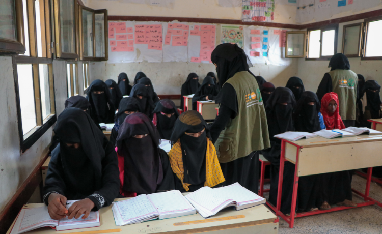 Two women in UNFPA vests and black head coverings walk among a classroom of girls with open text books on their desks