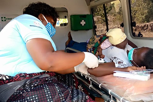 A health worker in a face mask inserts a contraceptive device into the arm of a woman holding a baby.