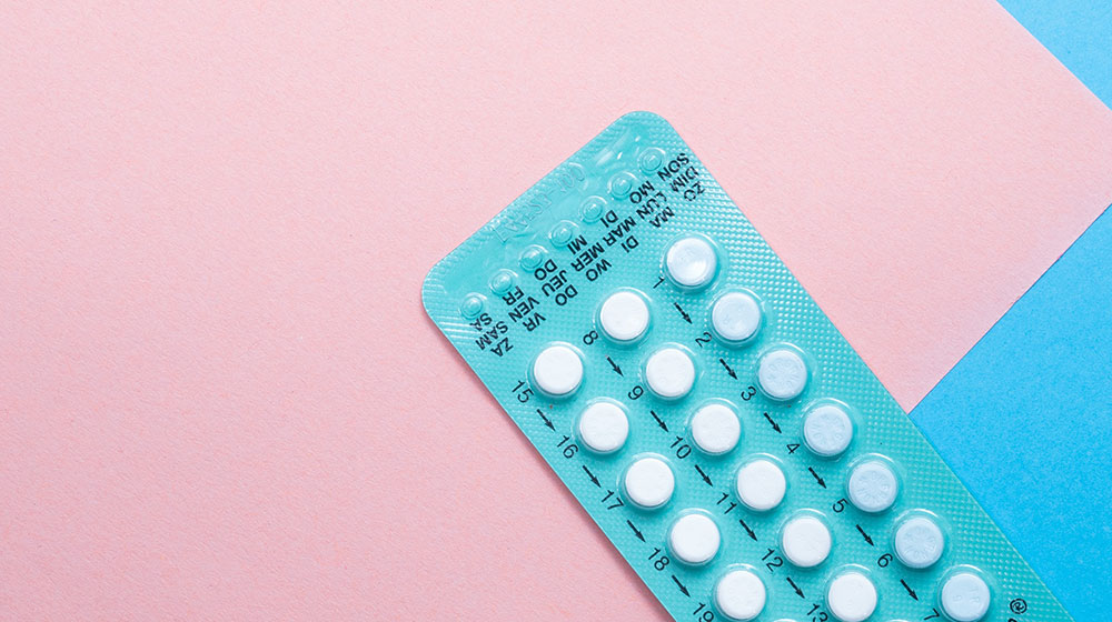 A photo shows a close-up of a pack of oral contraceptive pills on a pink and blue background.