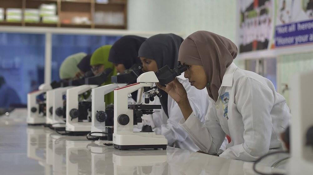 Mujeres con batas de laboratorio mirando al microscopio.