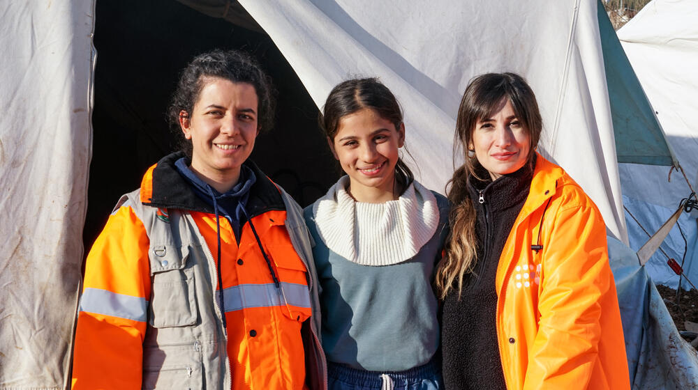 A young girl smiles and stands between two female humanitarian workers in front of a temporary camp in Türkiye. 