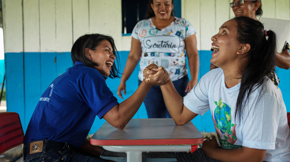 At an Indigenous school, two young women smile and hand wrestle as two older women look towards them and smile.