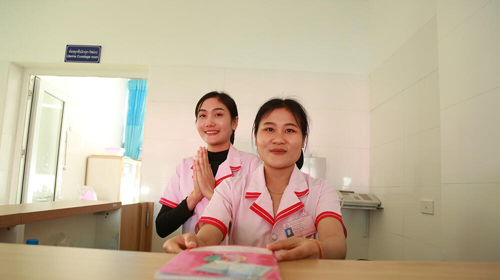 Two midwifery students sit behind a reception desk, one of them is clasping their hands.